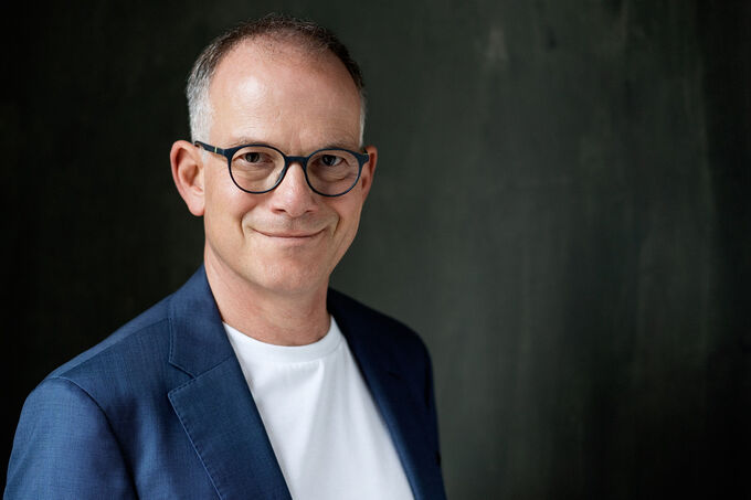 Ein lächelnder Mann mit Brille trägt einen blauen Blazer und ein weißes T-Shirt. Der Hintergrund ist dunkel und strukturiert. (Text KI generiert)