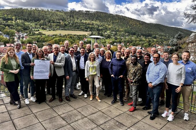Eine große Gruppe von Menschen steht im Freien, umgeben von einer grünen Landschaft und Bergen im Hintergrund. Einige Personen halten ein Dokument in der Mitte der Gruppe. Es ist sonnig und die Stimmung wirkt freundlich und einladend. (Text KI generiert)