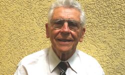 Ein älterer Mann mit grauen Haaren und Brille lächelt freundlich. Er trägt ein weißes Hemd und eine gestreifte Krawatte. Der Hintergrund ist gelb und strukturiert.