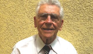 Ein älterer Mann mit grauen Haaren und Brille lächelt freundlich. Er trägt ein weißes Hemd und eine gestreifte Krawatte. Der Hintergrund ist gelb und strukturiert.