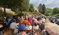 Eine große Gruppe von Menschen sitzt im Freien auf Stühlen, umgeben von Bäumen und Grünflächen. Einige Teilnehmer tragen Regenjacken und halten Regenschirme. Im Hintergrund sind Spielgeräte sichtbar. Die Atmosphäre wirkt entspannt und gesellig.