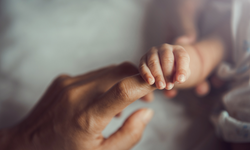 Eine Hand eines Erwachsenen berührt sanft die kleine Hand eines Babys. Der Fokus liegt auf den zarten Fingern des Babys und der Berührung, die eine liebevolle Verbindung symbolisiert. Der Hintergrund ist unscharf und neutral.