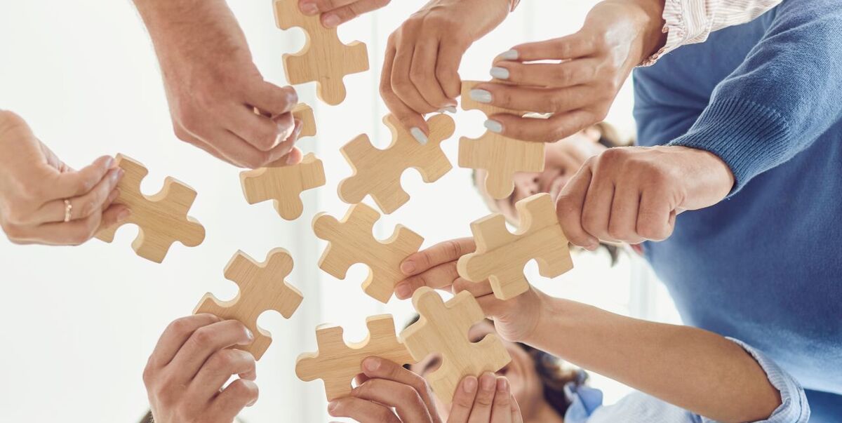 Mehrere Hände greifen nach Holzpuzzle-Teilen, die in der Luft schweben. Die Hände gehören verschiedenen Personen, die zusammenarbeiten, um das Puzzle zu lösen. Der Hintergrund ist hell und unscharf, was den Fokus auf die Hände und die Puzzlestücke lenkt. (Text KI generiert)