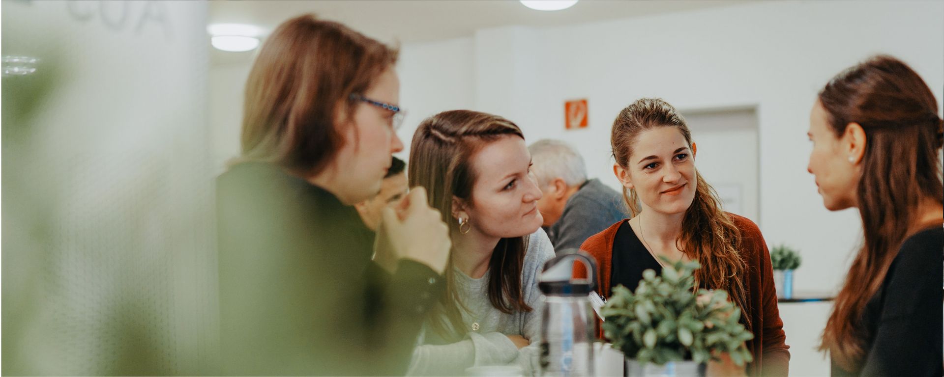 Vier Frauen sitzen an einem Tisch und unterhalten sich. Sie wirken interessiert und lachen. Im Hintergrund sind Pflanzen und eine Wasserflasche sichtbar. Die Atmosphäre ist freundlich und entspannt. (Text KI generiert)