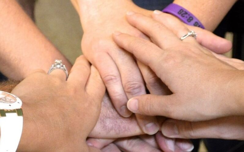Mehrere Hände liegen übereinander, die eine symbolische Geste der Einheit oder Zusammenarbeit darstellen. Einige Hände tragen Ringe und Armbänder. Der Hintergrund ist unscharf und zeigt keine spezifischen Details.