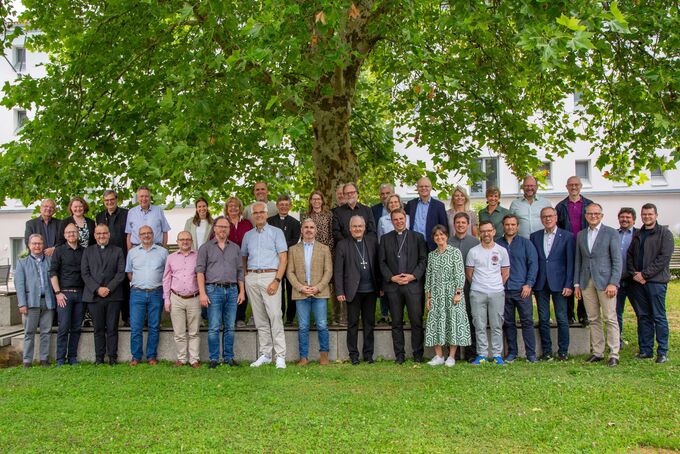 5. ökumenische Begegnung in Passau Eine große Gruppe von Menschen steht vor einem Baum auf einer Wiese. Die Personen sind in verschiedenen Kleidungsstilen gekleidet und lächeln in die Kamera. Es handelt sich um eine formelle Zusammenkunft oder ein Gruppenfoto. (Text KI generiert)