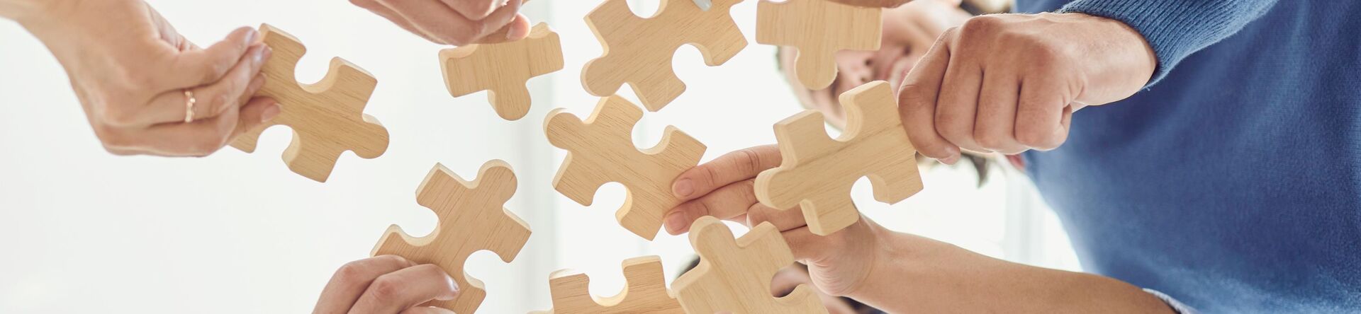 Mehrere Hände halten Holzpuzzle-Teile, die in die Luft gehalten werden. Die Hände gehören verschiedenen Personen, die gemeinsam an einem Puzzle arbeiten. Der Hintergrund ist hell und neutral. (Text KI generiert)