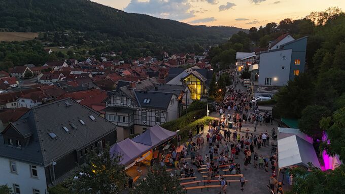 Eine belebte Straße in einer Stadt während eines Festes bei Sonnenuntergang. Menschen stehen zusammen, es gibt Stände und Zelte. Im Hintergrund sind Häuser und Hügel zu sehen, die eine malerische Landschaft bilden.