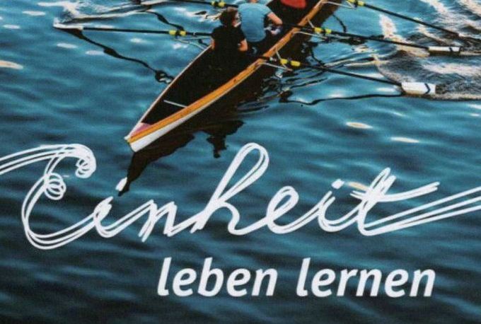 Zwei Personen rudern in einem Boot auf ruhigem Wasser. Der Text "Einheit leben lernen" ist prominent im Vordergrund platziert. (Text KI generiert)