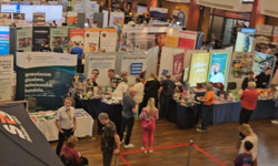 Eine Luftaufnahme einer Messehalle mit zahlreichen Ständen und Besuchern. Verschiedene Informationsstände sind zu sehen, die Produkte und Dienstleistungen präsentieren. Menschen bewegen sich zwischen den Ständen, während einige Gespräche führen. Ein großes Zelt mit dem Logo "SRS" ist im Vordergrund sichtbar.